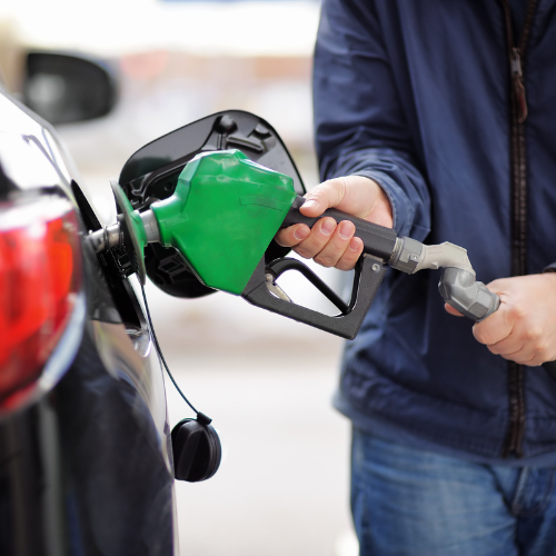 A person in a dark jacket refuels a car, inserting a green gas pump nozzle into the vehicle’s fuel tank. The scene conveys routine and focus.